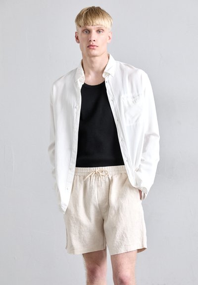 Chemise blanche à boutons au-dessus d'un débardeur noir côtelé, associée à un short en lin beige avec une taille élastique et un cordon de serrage.