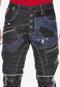 Blaue und schwarze Jeanshose mit mehreren Taschen, Metallnieten, dekorativen Nähten und einzigartigen Paneeldesigns mit einem Ledeelement.