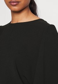 Svart blus med en slät textur, båthalsning och subtila puffärmar, med en enkel, minimalistisk design.