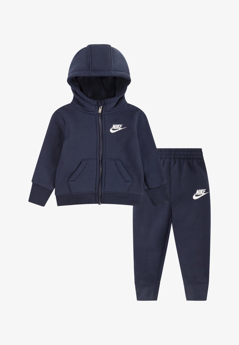 Hanorac albastru închis cu fermoar, glugă, buzunare frontale și logo alb Nike; asortat cu pantalonii sport strânși pe gleznă, cu detaliu de logo.