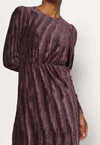 Mujer con un vestido largo de manga larga, plisado, de terciopelo color ciruela brillante, con una cintura ceñida, de pie contra un fondo blanco liso.
