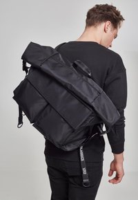 Sort rygsæk med en rulletopdesign, lavet af glat nylon. Har flere lynlåslommer og justerbare stropper.