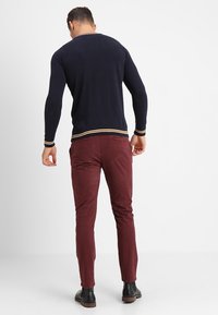 Maglione a maglia blu navy con polsini e orlo a righe, abbinato a pantaloni bordeaux slim-fit. Stivali in pelle nera completano l'outfit.
