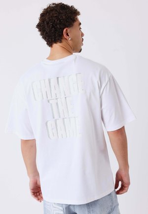 Uomo con capelli ricci che indossa una maglietta bianca con il testo "CHANGE THE GAME" sulla schiena, guarda di lato, abbinata a jeans di denim chiaro.