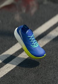 Zapatilla de correr azul con un upper texturizado, cordones azul claros y una suela amarilla. Presenta un diseño elegante con orificios de ventilación y una forma aerodinámica.