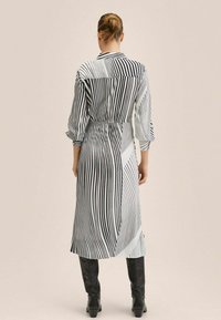 Femme avec les cheveux en chignon, portant une robe midi rayée verticalement noir et blanc et des bottes noires montantes, se tenant de dos.