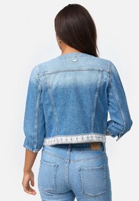 Chaqueta de mezclilla, azul claro, diseño corto con mangas de tres cuartos. Con un dobladillo de encaje, detalles de costura sutiles y una etiqueta de logo en la parte trasera.