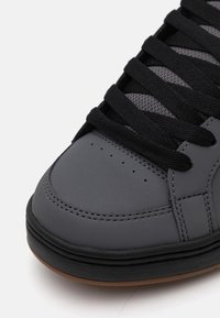 Sneaker grigio con una tomaia in pelle liscia e mesh, dotato di lacci neri, dettagli perforati e una suola in gomma testurizzata.