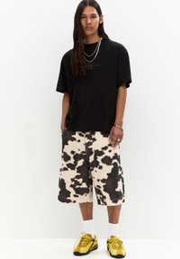 T-shirt nera oversize, pantaloni corti a stampa mucca e sneakers gialle con calze bianche. Dettagli notevoli includono collane a strati e una vestibilità rilassata.