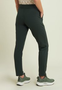 Pantaloni slim-fit verde scuro con una texture liscia, vita elasticizzata, tasche laterali, abbinati a scarpe da ginnastica grigio chiaro.