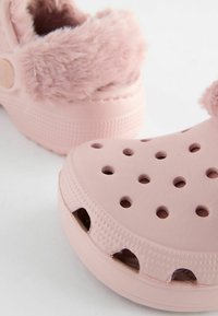Sabots rose pâle avec doublure douce, présentant plusieurs trous de ventilation, une texture à motifs sur les côtés et une forme arrondie.
