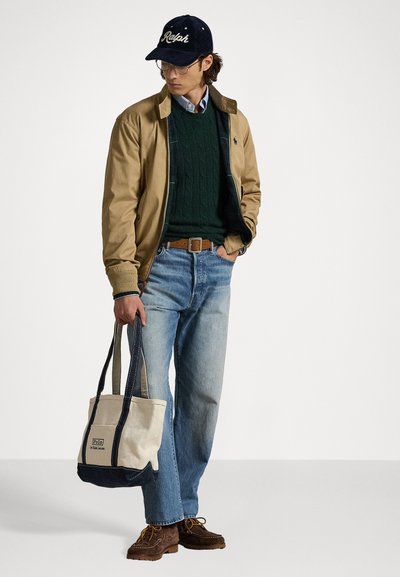Braune Bomberjacke über einem grünen Strickpullover, blaue Jeans, braune Schnürschuhe und eine beige Tote-Tasche mit blauen Akzenten. Marineblauer Hut mit Logo.