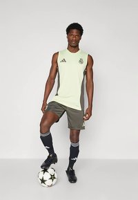 Camisola de futebol verde sem mangas com painéis laterais cinzentos, usada com calções escuros, chuteiras pretas e meias pretas às riscas, em pé ao lado de uma bola de futebol.