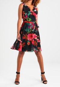 Czarna sukienka w kwiaty z żywymi różowymi i zielonymi wzorami, bez rękawów, z dopasowaną talią i rozkloszowanym dołem. Czarne sandały na obcasie.