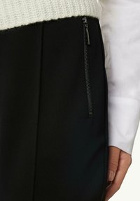 Czarna spódnica z materiału posiada boczny zamek błyskawiczny i plisy, zestawiona z białym, teksturowanym swetrem w okolicy talii. Zbliżenie na materiały.