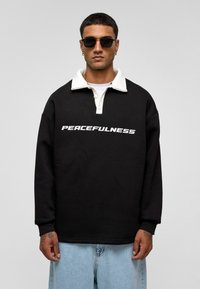 Zwarte sweatshirt met een witte kraag en de tekst "PEACEFULNESS". Gemaakt van een zachte stof, met lange mouwen en een relaxte pasvorm.