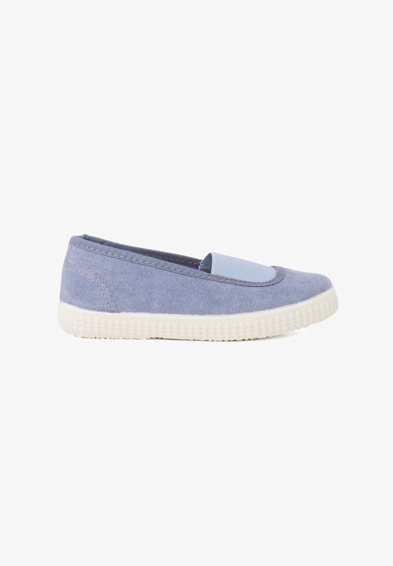 Des chaussures slip-on en toile bleu clair avec une semelle en caoutchouc blanc texturé. Elles présentent un dessus lisse avec des coutures et un accent bleu uni.