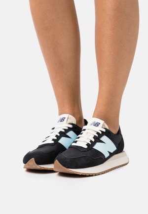 Personne portant des baskets New Balance noires et bleu clair avec des lacets blancs et des semelles beiges, debout sur une surface blanche.