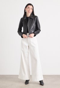 Vrouw staat in een studio en draagt een zwarte leren jas, een zwarte top en brede witte broek, met lang haar en opvallende oorbellen.