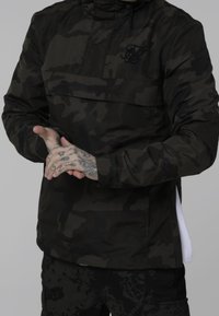 Person iført sort og mørkegrå camouflaget jakke og bukser med tatoverede hænder sammenfoldet foran.