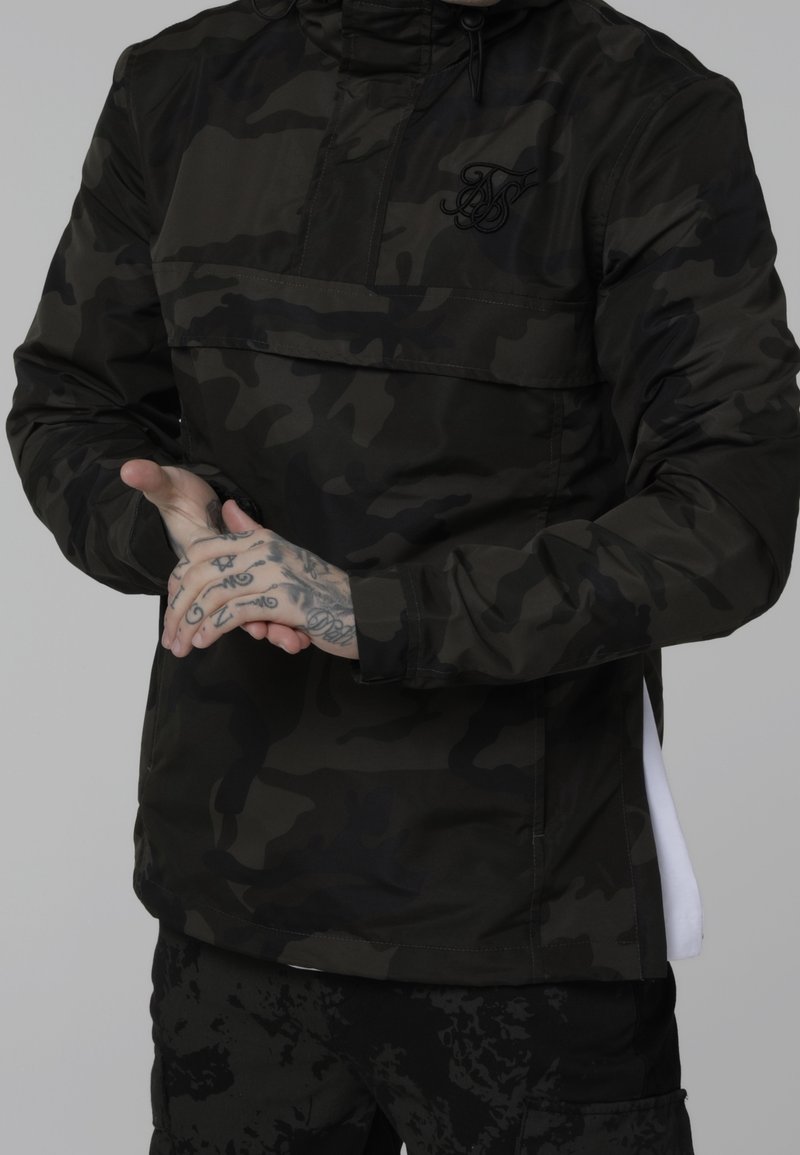 Person iført sort og mørkegrå camouflaget jakke og bukser med tatoverede hænder sammenfoldet foran.