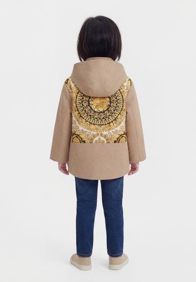 Olkapäihin ulottuvat tummat hiukset omaava lapsi, jolla on päällään beige hupullinen takki, jonka selkä on koristeltu koristeellisella kultaisella ja mustalla kuviolla, siniset farkut ja beigejä kenkiä, seisomassa.