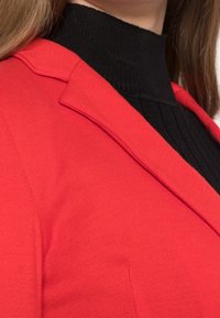 Gros plan sur une personne portant un blazer rouge texturé par-dessus un pull à col roulé noir côtelé, avec des cheveux raides châtain clair.