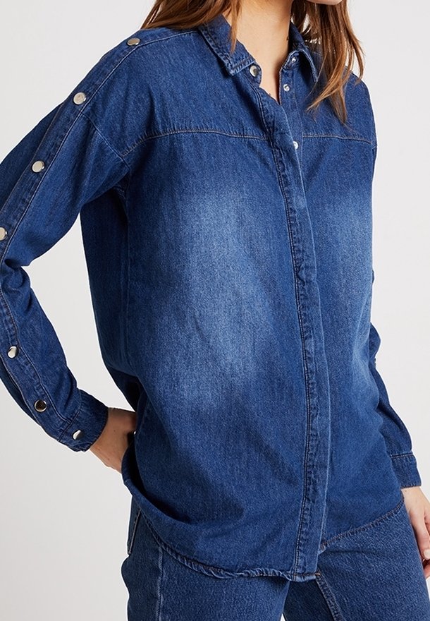 Femme portant une chemise en denim bleu avec des boutons-pression métalliques le long des manches et à l'avant, assortie à un jean en denim.