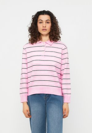 Femme aux cheveux bouclés portant un pull rose à col avec des rayures horizontales noires et blanches, et un jean bleu, debout devant un fond uni.