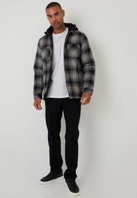 Schwarz-graue karierte Flanelljacke mit Kapuze, zwei Brusttaschen und einem weichen Innenfutter. Kombiniert mit einem weißen T-Shirt und schwarzen Hosen.