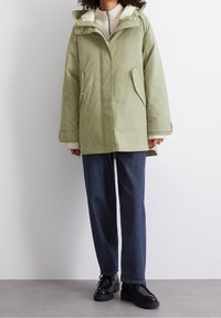 Grön isolerad jacka med fuskpälsfodrad huva, dragkedjestängning, två sidofickor och breda ärmar. Bärs med mörka denimbyxor och svarta skor.