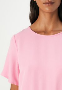 Chemise rose à manches courtes avec un col rond, fabriquée à partir d'un tissu lisse et texturé. Couture visible le long du col et des manches.