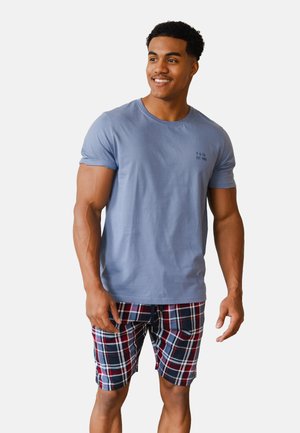 Ein lächelnder Mann steht vor einem einfarbigen Hintergrund und trägt ein hellblaues T-Shirt mit kurzen Ärmeln sowie rot-weiß-blaue karierte Shorts.