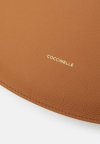 Tan leather item with a textured finish, featuring a gold "Coccinelle" logo, and visible stitching along the edge.