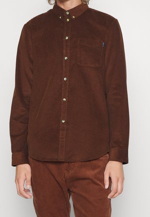 Uomo che indossa una camicia marrone in velluto a coste a maniche lunghe con bottoni e pantaloni coordinati in velluto a coste marrone su uno sfondo semplice.