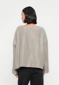 Luźny, szary top wykonany z miękkiej tkaniny. Posiada dekolt typu łódka i długie rękawy, z nieco dłuższym tylnym brzegiem. Czysty, prosty design.