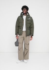 Groene pufferjack met capuchon, ritssluiting en voorzakken; gecombineerd met een lichtgrijs t-shirt en beige wijde broek. Witte sneakers en tas.
