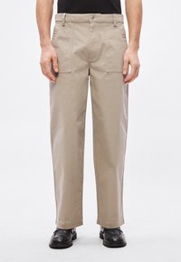 Bézs színű, pamut egyenes szárú nadrág oldal-zsebekkel és egyszerű gombos záródással. Fekete cipővel viselve, egyszínű háttér előtt.