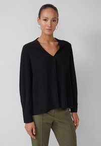 Schwarz, langärmlige Bluse mit V-Ausschnitt und dezenten Falten, aus weichem Stoff gefertigt. Kombiniert mit olivgrünen, maßgeschneiderten Hosen.