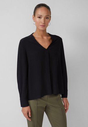 Schwarz, langärmlige Bluse mit V-Ausschnitt und dezenten Falten, aus weichem Stoff gefertigt. Kombiniert mit olivgrünen, maßgeschneiderten Hosen.