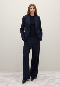 Completo su misura blu navy composto da blazer monopetto e pantaloni a gamba larga, realizzato in tessuto liscio con una lieve lucentezza. Top nero sotto.