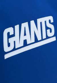 Niebieska odzież z napisem "GIANTS" w dużych, pogrubionych białych haftowanych literach z poziomą linią pod spodem. Gładka tekstura materiału.