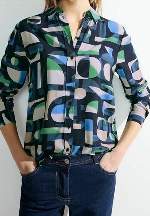 Frau trägt ein langärmliges Hemd mit abstraktem geometrischem Muster in Blau, Grün, Schwarz und Beige, in dunkelblaue Jeans gesteckt.