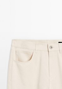 Beige denim shorts, featuring a front pocket, metal button closure, and belt loops. Textured fabric with a smooth finish.