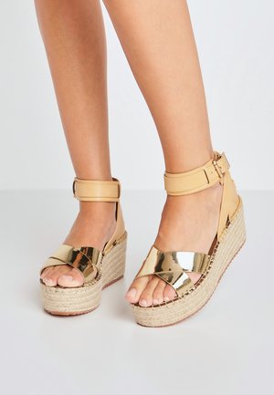 Gold metallic wedge sandals with cross-strap design, natural woven jute platform, and adjustable tan ankle strap. Smooth texture.