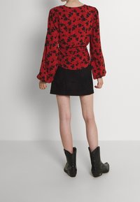 Blouse rouge à manches longues avec motif floral noir, associée à une mini-jupe en daim noir et des bottes de cow-boy noires. Vue de dos présentée.