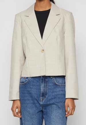 Vrouw draagt een lichtbeige enkelknopsblazer over een zwarte top en blauwe spijkerbroek, getoond van schouders tot heupen.