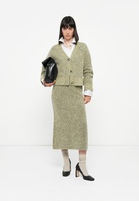 Green textured knit cardigan and matching skirt, white collared shirt underneath, holding a black leather bag, paired with black high heels.