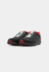 Black athletic shoes with red accents, grey laces, mesh tongue, and textured black outsole featuring red patterns. Three black stripes on sides.