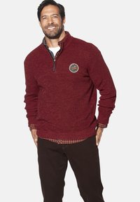 Roter strukturierten Pullover mit einem Viertel-Reißverschluss, versehen mit einem Logo-Patch. Über einem karierten Hemd getragen, kombiniert mit dunklen Hosen.
